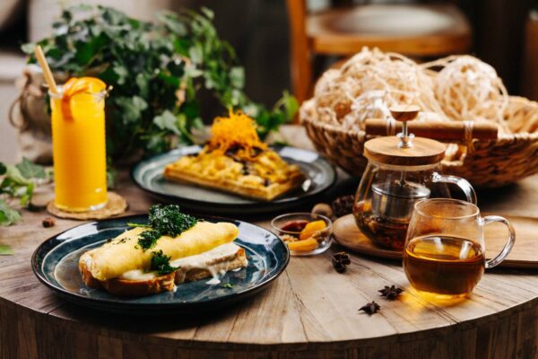 A rustic breakfast spread featuring an omelette on toast, a plate of waffles topped with shredded garnish, a glass of orange juice, and a teapot with a matching cup of herbal tea. The dishes are arranged on a wooden table with a basket of bread and greenery in the background, creating a warm and inviting morning atmosphere.