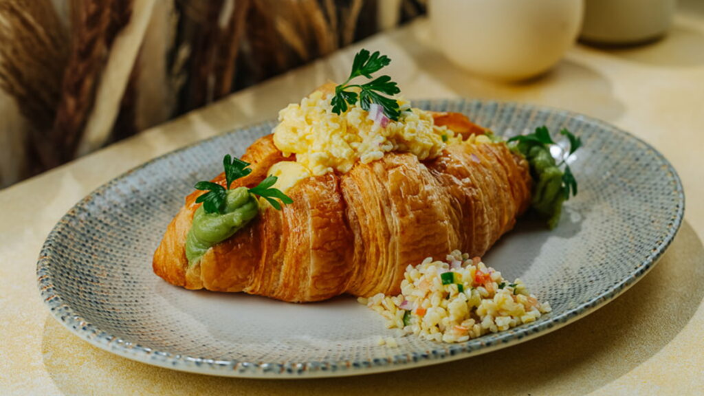 Croissant Scramble: Homemade Croissant bread with scrambled egg, smashed avocado, tomato, beef, chives, parsley, mint, cucumber, onion, and spring onion.