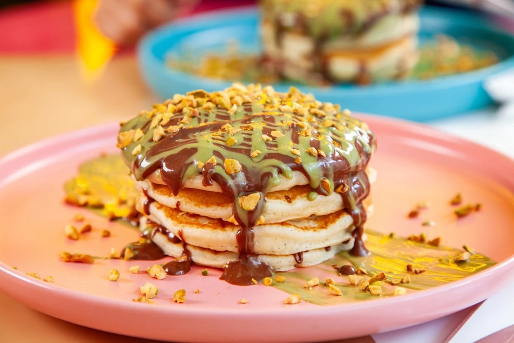Stack of fluffy pancakes topped with chocolate and pistachio sauce, sprinkled with crushed nuts, served on a pastel plate at BohoX café.