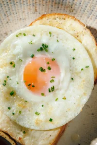 Close-up of a sunny-side-up egg on toasted bread, topped with chopped herbs, showcasing a simple and fresh breakfast dish