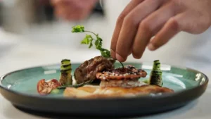Chef plating a fine dining dish with grilled meat, sesame-crusted seafood, and garnish.