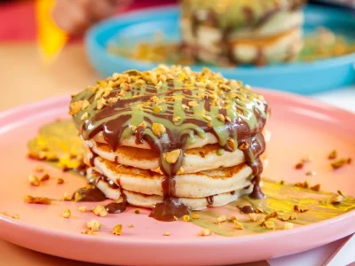 Stack of fluffy pancakes topped with chocolate and pistachio sauce, sprinkled with crushed nuts, served on a pastel plate at BohoX café.