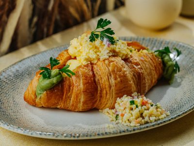Croissant Scramble: Homemade Croissant bread with scrambled egg, smashed avocado, tomato, beef, chives, parsley, mint, cucumber, onion, and spring onion.