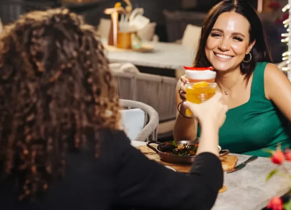 Two women sitting at a restaurant table, smiling and raising their drinks in a cheerful toast. One woman faces the camera holding a yellow beverage, while the other, in a green top, smiles warmly while holding a layered drink with a red garnish. A bowl of food sits on the table between them, creating a cozy and friendly dining atmosphere.