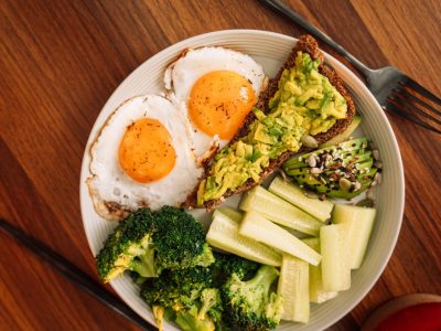 Healthy breakfast plate with two sunny side up eggs, avocado toast, sliced cucumber, steamed broccoli, and avocado topped with seeds on a wooden table.