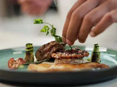 Chef plating a fine dining dish with grilled meat, sesame-crusted seafood, and garnish.